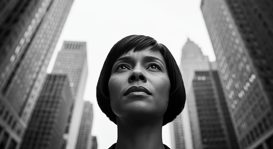 A low-angle close-up shot, in stark black and white, focuses on a woman with a short, precisely cut bob. Her expression is one of deep concern; her eyebrows are slightly furrowed, her mouth drawn into a thin line, and her eyes hold a worried intensity. The high contrast of the black and white photography emphasizes the texture of her skin and the lines around her eyes, accentuating her worried expression. The background is a blurred but imposing array of tall skyscrapers, their forms rendered in varying shades of grey, creating a sense of depth and scale. The low angle, shooting upwards, emphasizes her upward gaze, suggesting a sense of being overwhelmed by the weight of her worries within the vast urban landscape. The overall mood is one of serious apprehension, a powerful and poignant image of a woman grappling with anxieties within a monumental city.
